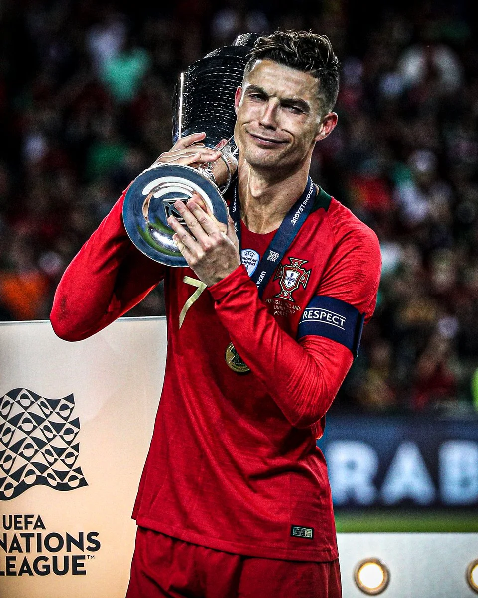 Cristiano Ronaldo in a red Portugal jersey holding the UEFA Nations League trophy with a playful expression.